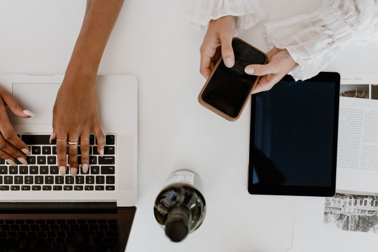Overhead picture of what appears to be a white desk with black female hands typing on a laptop and white female hands holding a cell phone. A black iPad or tablet is laying on the desk.