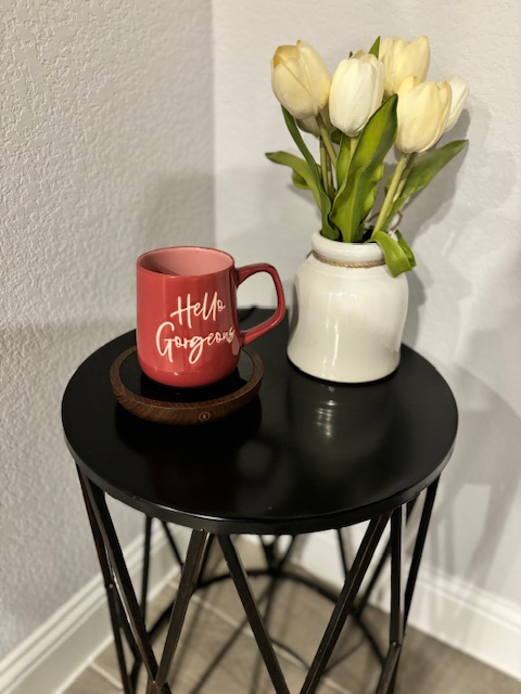 Black table with a white vase and white flowers. Sweetfull coffee mug warmer with a pick coffee mug is also on the table.
