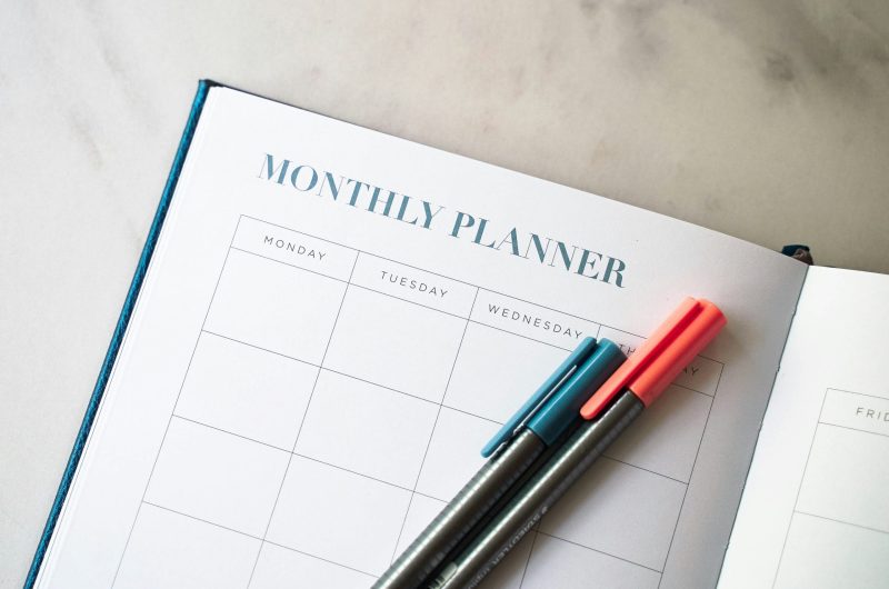 Open monthly planner with a blank calendar layout on a marble surface, featuring two pens in teal and coral resting across the page.