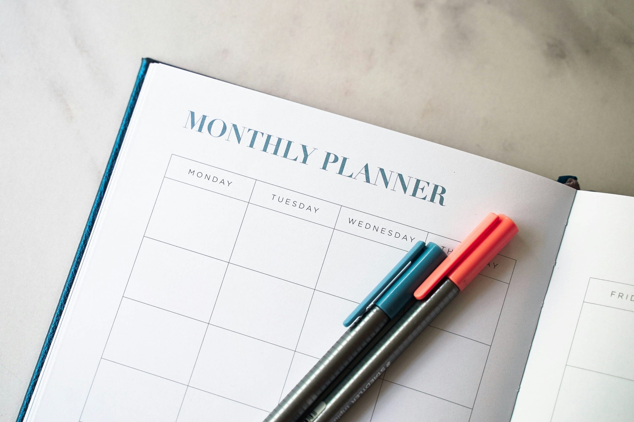 Open monthly planner with a blank calendar layout on a marble surface, featuring two pens in teal and coral resting across the page.