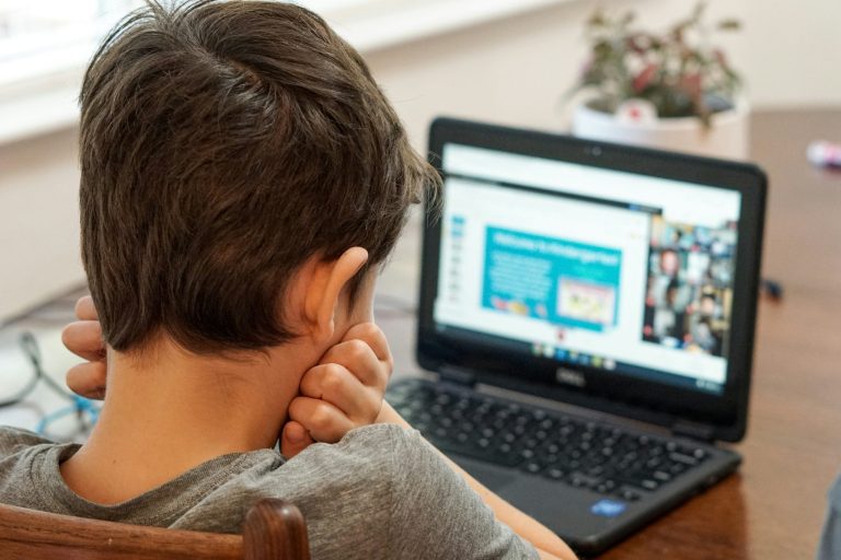 Child sitting at a table with head resting on hands, looking at a laptop screen displaying an online class or video call.