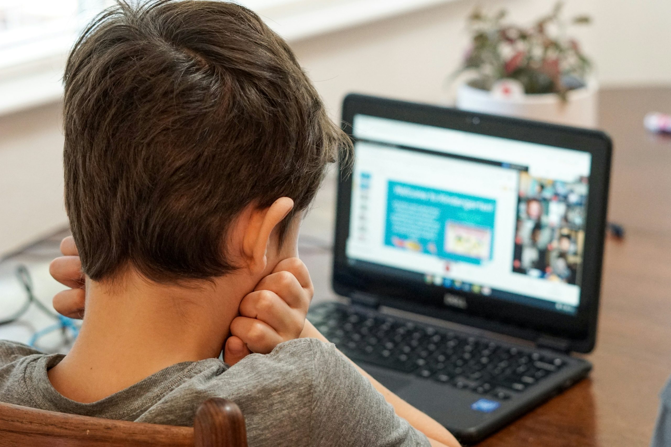 Child sitting at a table with head resting on hands, looking at a laptop screen displaying an online class or video call.