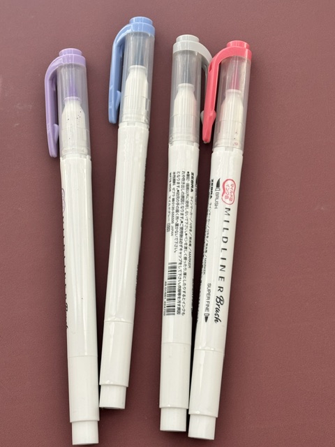 Four pastel Zebra Mildliner brush markers in purple, blue, gray, and pink on a desk.