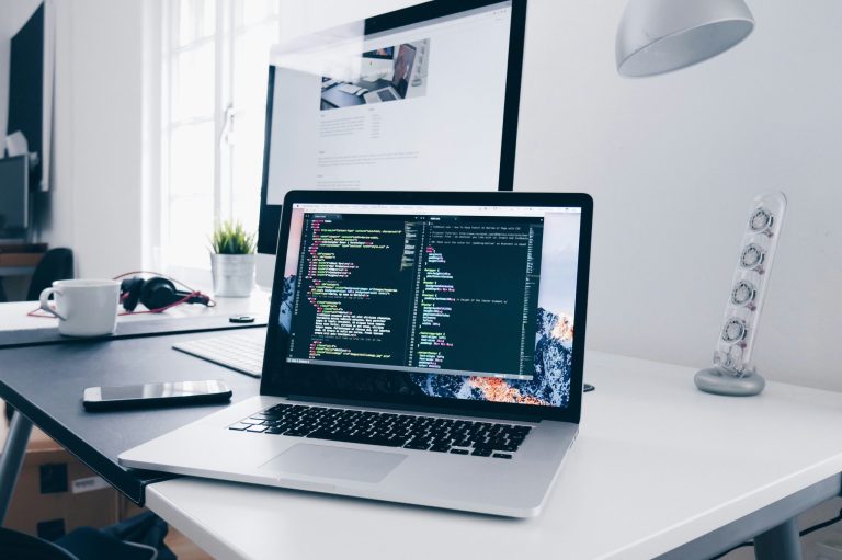 Modern workspace featuring a laptop displaying website code, an external monitor, and office accessories on a white desk, illustrating website development and the importance of online privacy tools such as CookieYes.