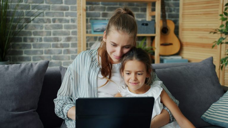 Mother and young daughter sitting together on a gray couch, smiling as they look at a laptop screen in their living room.